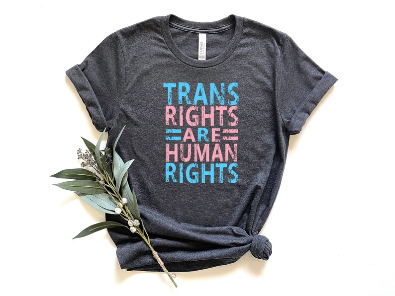 May include: A dark heather grey t-shirt with the text "TRANS RIGHTS ARE HUMAN RIGHTS" printed in pink and blue.