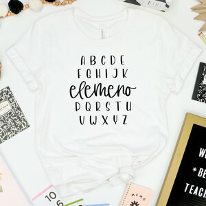 以下が含まれることがあります： 白いTシャツに黒い文字で「ABCDE FGHIJK elemeno PQRSTU VWXYZ」と書いてあります。Tシャツは白い表面に置かれており、その周りに黒と白のノート、金色の文字盤に「WORLDS *BEST* TEACHER」と書かれたもの、クレヨンなど、学校の用品が置かれています。