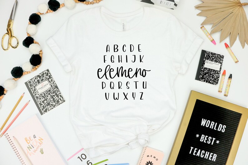 以下が含まれることがあります： 白いTシャツに黒い文字で「ABCDE FGHIJK elemeno PQRSTU VWXYZ」と書いてあります。Tシャツは白い表面に置かれており、その周りに黒と白のノート、金色の文字盤に「WORLDS *BEST* TEACHER」と書かれたもの、クレヨンなど、学校の用品が置かれています。