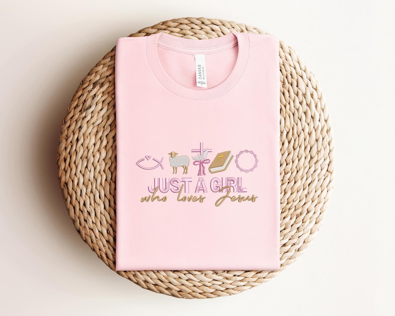 May include: Pink t-shirt with a graphic design of religious symbols and the text "JUST A GIRL who loves Jesus".