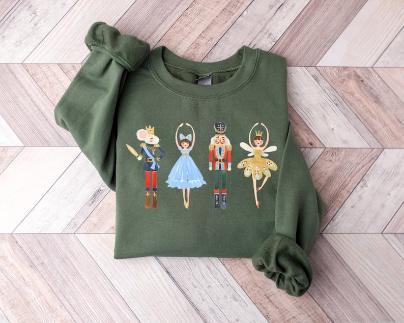 May include: A green sweatshirt with a festive design featuring a mouse, two ballerinas, and a nutcracker. The ballerinas are wearing blue and gold tutus.
