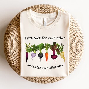 May include: A white t-shirt with a graphic of colorful root vegetables and the text "Let's root for each other and watch each other grow".