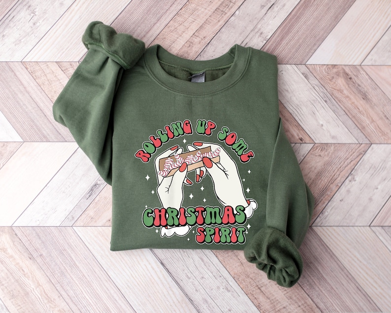 Puede incluir: Sudadera verde con un gr&aacute;fico blanco de manos que enrollan un porro con el texto "Rolling Up Some Christmas Spirit" en rojo, verde y blanco.