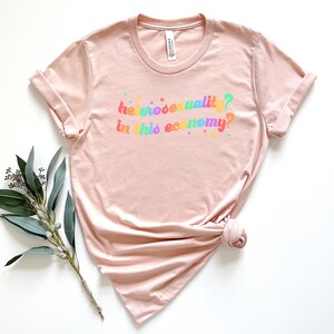 Pu&ograve; includere: Una maglietta rosa chiaro con la scritta "eterosessualit&agrave;? in questa economia?" stampata in colori arcobaleno.