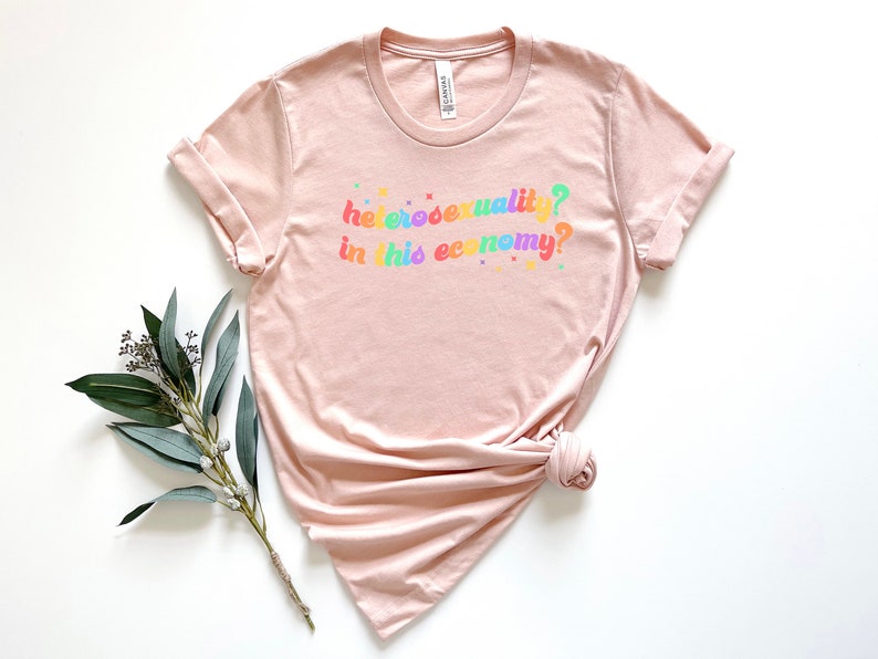 Pu&ograve; includere: Una maglietta rosa chiaro con la scritta "eterosessualit&agrave;? in questa economia?" stampata in colori arcobaleno.