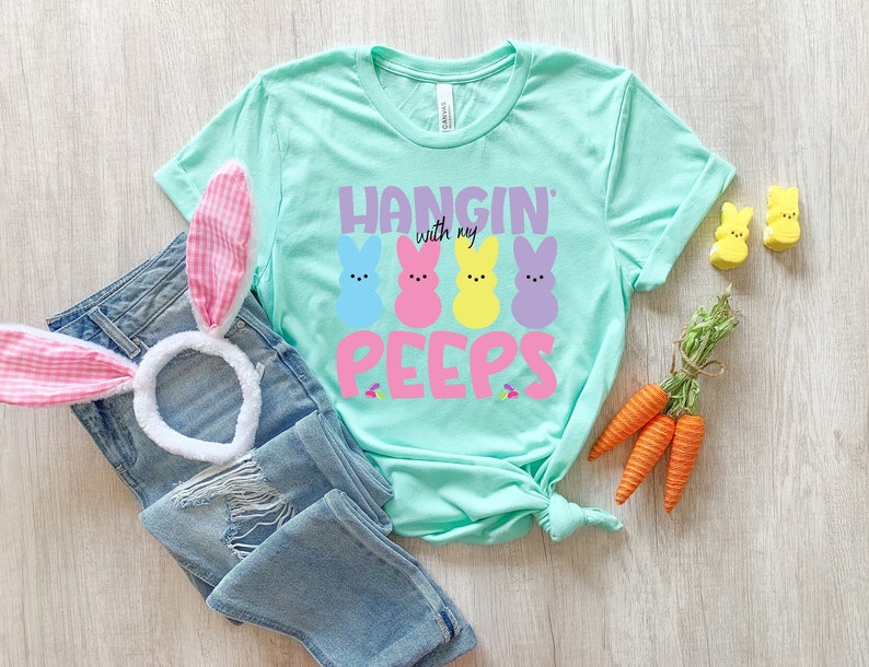 May include: A turquoise blue t-shirt with the text "Hangin' with my Peeps" in pink, purple, and yellow letters. Four colorful marshmallow Peeps are pictured above the text. The shirt is laid out on a wooden surface with a pair of pink and white bunny ears and a pair of blue jeans.