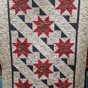 Puede incluir: Una colcha roja y blanca con un patrón geométrico en blanco y negro. La colcha presenta un diseño de estrella repetido con las palabras "Peace", "Joy", "Christmas" y "Noel" escritas dentro de cada estrella.