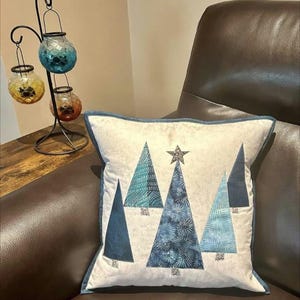 Könnte beinhalten: Ein weißes Kissen mit einem blauen und weißen Patchwork-Muster aus fünf stilisierten Weihnachtsbäumen. Das Kissen liegt auf einem braunen Ledersofa.