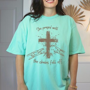 May include: A light turquoise t-shirt with the text "She prayed until the chains fell off" in brown. The design includes a brown cross and broken chains.