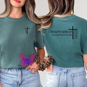 Op de afbeelding: Een tealblauw t-shirt met een kruis en de tekst "GRACE" op de voorkant en de tekst "I THROW MY HANDS UP AND PRAISE YOU AGAIN AND AGAIN" op de achterkant. Het shirt wordt gedragen door een persoon die blauwe jeans draagt.