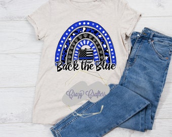 Peut inclure: Un t-shirt beige clair avec un motif arc-en-ciel comportant un badge de police au centre. L'arc-en-ciel est composé de différents motifs, dont l'imprimé léopard, les pois et les cœurs. Le texte "Back the Blue" est imprimé sous l'arc-en-ciel.