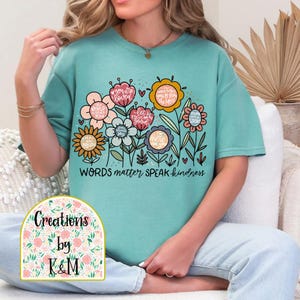 May include: A turquoise blue t-shirt with a floral design and the text "WORDS matter SPEAK kindness". The flowers are pink, yellow, and green with various positive messages written on the petals. The t-shirt is a relaxed fit and has short sleeves.