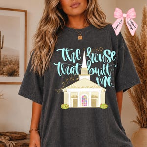 May include: A charcoal grey short-sleeve t-shirt featuring a graphic of a church building with a steeple and musical notes. The text reads "the house that built me" in light blue script. A pink bow graphic is positioned in the upper right corner.