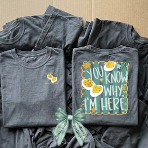 May include: A charcoal gray t-shirt featuring a small graphic of deviled eggs on the left chest and a larger matching graphic on the back with the text "YOU KNOW WHY I'M HERE" surrounded by a yellow scalloped border and teal vertical stripes.