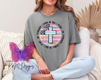 May include: Gray short-sleeved t-shirt featuring a circular design with an American flag and a cross. The text "on freedom I stand, in God I trust" surrounds the design. The shirt is worn by a person.