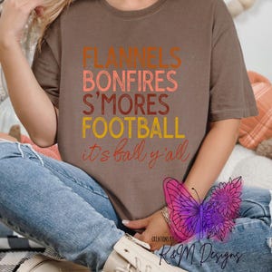 May include: A brown t-shirt with the text "Flannels, Bonfires, S'mores, Football, it's fall y'all" in orange, red, and yellow. The text is stacked vertically. The shirt is being worn by a person. The image also includes a butterfly and sunflower graphic.