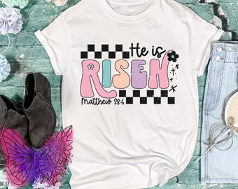 May include: White t-shirt with a colorful graphic that says "He is Risen" with a checkered pattern and the text "Matthew 28:6".