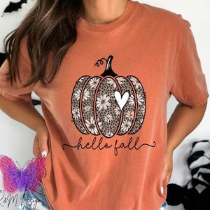 May include: A burnt orange t-shirt with a graphic of a pumpkin with a white heart and floral pattern. The text "hello fall" is written below the pumpkin.