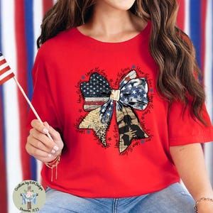 May include: A red t-shirt featuring a patriotic bow design. The bow incorporates the American flag, camouflage, and text that reads "Honor the Brave." The shirt is a casual style, suitable for patriotic events.