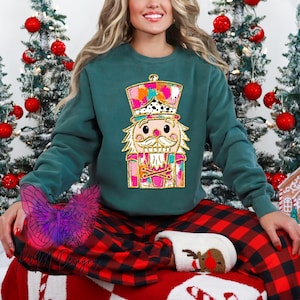 May include: A teal blue sweatshirt with a colorful nutcracker design. The nutcracker is wearing a pink and gold hat and has a pink and gold beard. The sweatshirt is paired with red and black plaid pajama pants.