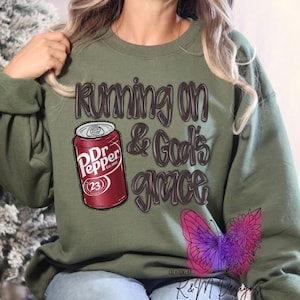 May include: Green sweatshirt with the text "Running on & God's grace" and a red and white Dr Pepper can graphic. The sweatshirt also features a purple butterfly graphic.