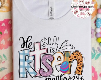 May include: White t-shirt with a colorful graphic design that says "He is risen" with a cross and an Easter egg. The text "matthew 28:6" is below the graphic.