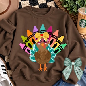 May include: A brown crewneck sweatshirt featuring a graphic of a turkey with colorful crayon tail feathers, wearing pink glasses and a yellow bow. A separate light green ribbon with the text Roman's 8:28 and K&M Designs is attached.