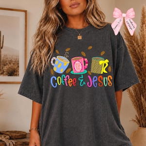 May include: A charcoal gray short-sleeve t-shirt featuring a graphic of three coffee mugs and the text "Coffee & Jesus". The mugs are blue with yellow spots, pink with the text "Jesus is my fuel", and leopard print with a yellow bow.
