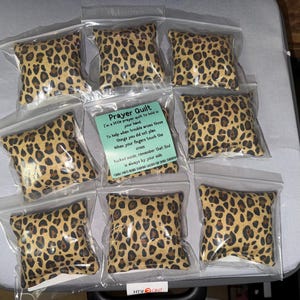 May include: A collection of small, square prayer quilts in clear plastic bags. The quilts feature a tan and brown leopard print pattern. A small card with text about the prayer quilt is visible in one of the bags.