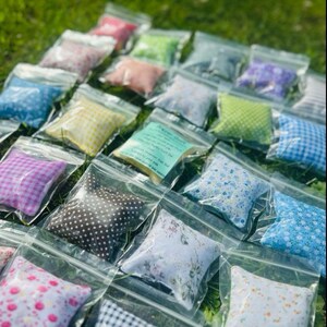 May include: An assortment of small, colorful fabric pillows in clear plastic bags. The pillows feature various patterns, including polka dots, floral prints, and gingham checks. Some bags contain a small card with text. The pillows are arranged on a bed of green grass.