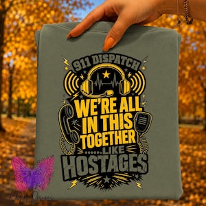 May include: An olive green t-shirt featuring a graphic design with yellow and black text that reads 911 DISPATCH WE'RE ALL IN THIS TOGETHER LIKE HOSTAGES. The design includes illustrations of a headset, a telephone receiver, and a radio microphone.