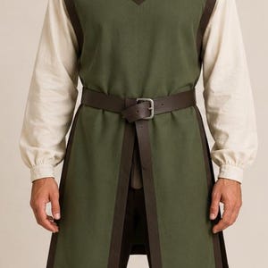 May include: A sleeveless olive green tunic featuring dark brown trim along the edges and a notched neckline. The tunic is worn over a long-sleeved cream-colored shirt and is cinched at the waist with a dark brown leather belt with a metal buckle.