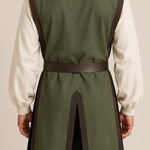 May include: A sleeveless olive green tunic with dark brown trim along the edges, worn over a long-sleeved cream-colored shirt. A dark brown belt is cinched at the waist. The tunic features a split back hem and is paired with dark brown trousers.