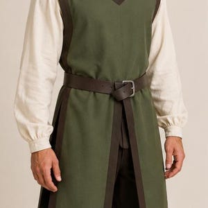 May include: An olive green sleeveless tunic featuring dark brown trim along the edges, a notched neckline, and side slits. The tunic is worn over a long-sleeved cream-colored shirt and dark brown trousers, cinched at the waist with a brown leather belt.