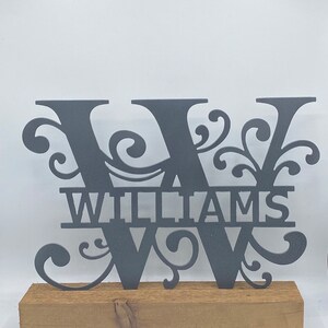 May include: A dark gray metal monogram with the letter "W" and the name "WILLIAMS" cut out. The monogram sits on a rectangular wooden base. The design includes decorative flourishes.