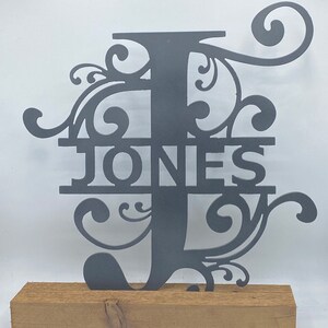 Puede incluir: Un monograma de metal gris oscuro con la letra "J" y el nombre "JONES" recortados. El monograma presenta adornos decorativos y está montado sobre una base de madera natural. El fondo es blanco.