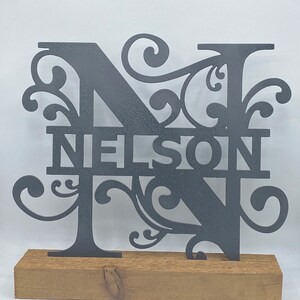 May include: A gray metal sign with the name "NELSON" cut out, featuring an ornate "N" and "T" with decorative flourishes. The sign is mounted on a rectangular, natural wood base, suitable for display.