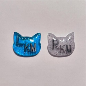 Può includere: Due spille in resina a forma di gatto. Una è blu vibrante con glitter e le lettere "LKM" in argento. L'altra è grigia traslucida con glitter argentati e le lettere "RKM". Entrambe le spille hanno una finitura lucida.