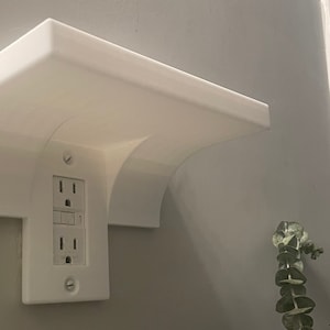 Könnte beinhalten: Ein weißes Regal mit einem geschwungenen Design, das an einer Wand neben einer weißen Steckdose mit zwei Steckplätzen montiert ist. Das Regal besteht aus einem glatten, weißen Material und hat eine leicht abgerundete Kante.