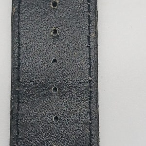 May include: A close-up of a black leather watch strap with four holes for adjusting the size.