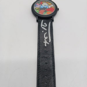 May include: A black leather watch band with a white signature that reads "Romero Britto". The watch face is not visible in the image.