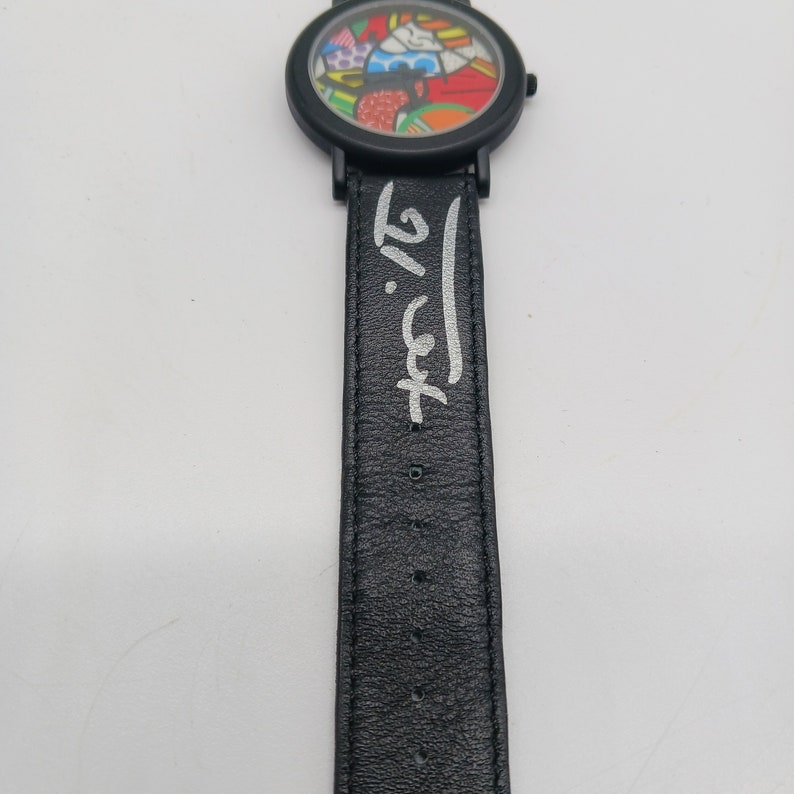May include: A black leather watch band with a white signature that reads "Romero Britto". The watch face is not visible in the image.