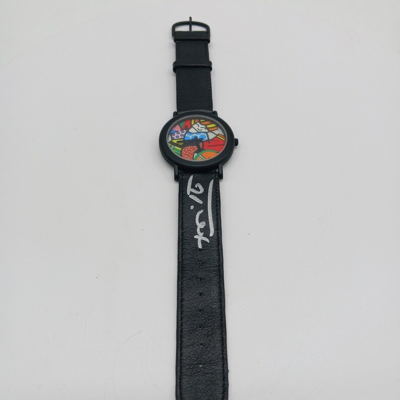 May include: A black wristwatch with a colorful, abstract design on the face. The watch band has the artist's signature, "K. Haring", in white lettering.