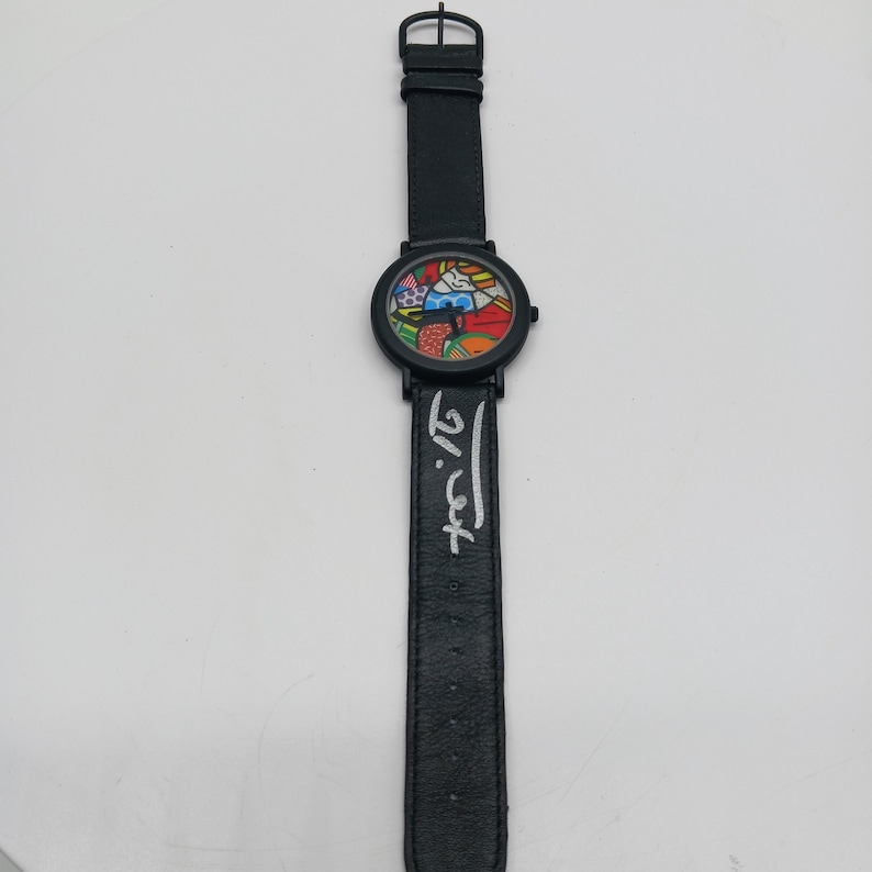 May include: A black wristwatch with a colorful, abstract design on the face. The watch band is black leather with the artist's signature in white lettering.