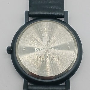 May include: Black wristwatch with a silver face and black leather band. The watch face has the text "R.T. 144/350" printed on it.