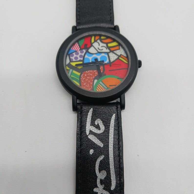 May include: Black wristwatch with a black leather band and a colorful, abstract face. The band has a white signature on it.