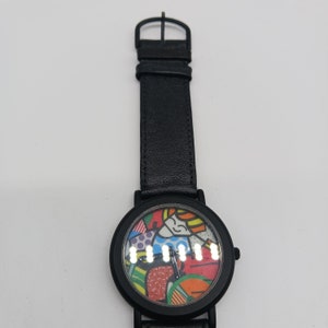 May include: Black leather wristwatch with a colorful abstract design on the face. The watch has a black leather band and a silver buckle.