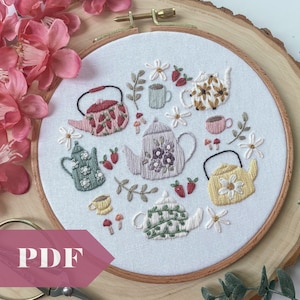 Könnte beinhalten: Stickerei in einem runden Rahmen mit einer verspielten Teeparty-Szene mit sechs Teekannen, Blumen und Erdbeeren. Die Teekannen sind in verschiedenen Farben und Mustern gehalten, darunter Grün, Blau, Rot und Gelb.