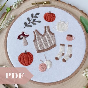 Può includere: Arte di ricamo a cerchio con un tema autunnale accogliente. Presenta un gilet a righe marroni e bianche, due zucche arancioni, un fungo, una teiera, una tazza di caffè, un gomitolo di lana e due calzini bianchi.