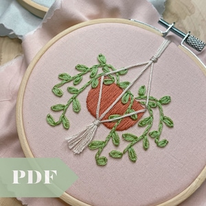 Op de afbeelding: Een close-up van een handgestikte borduurring met een hangend plantenontwerp. De plant is groen met een terracotta pot en een witte kwast. De ring bevindt zich op een roze stoffen achtergrond.